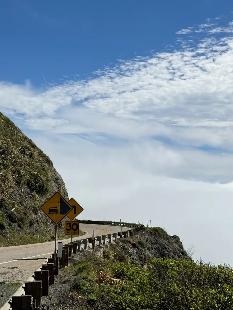 A curving coastal road above the fog with a caution sign